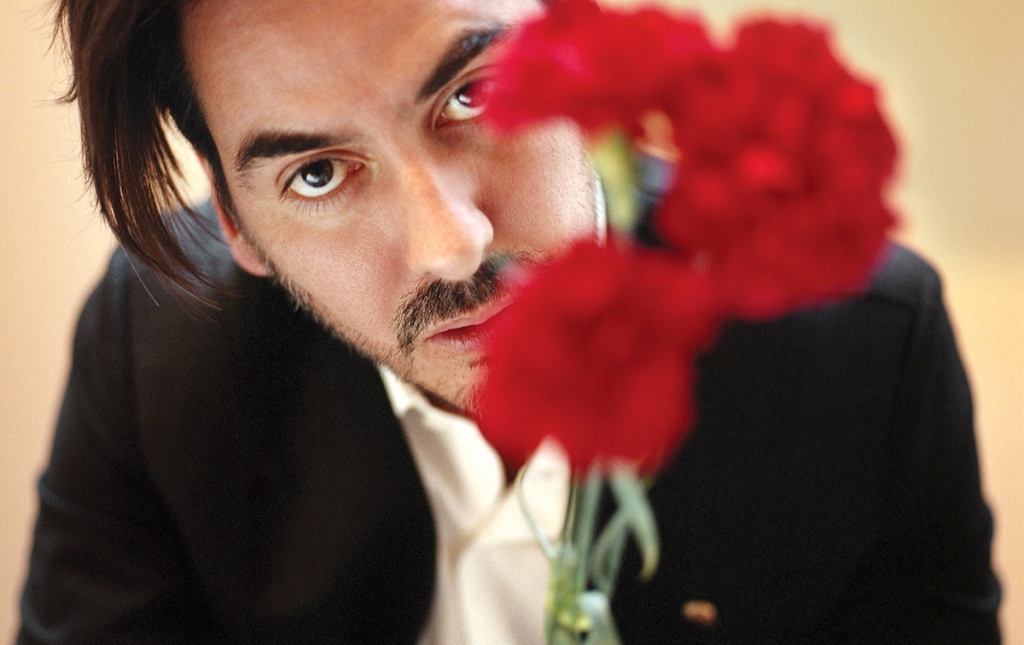 Image of Dhani Harrison ’01 in a black suit and white shirt, face partially obscured behind bright red flowers.
