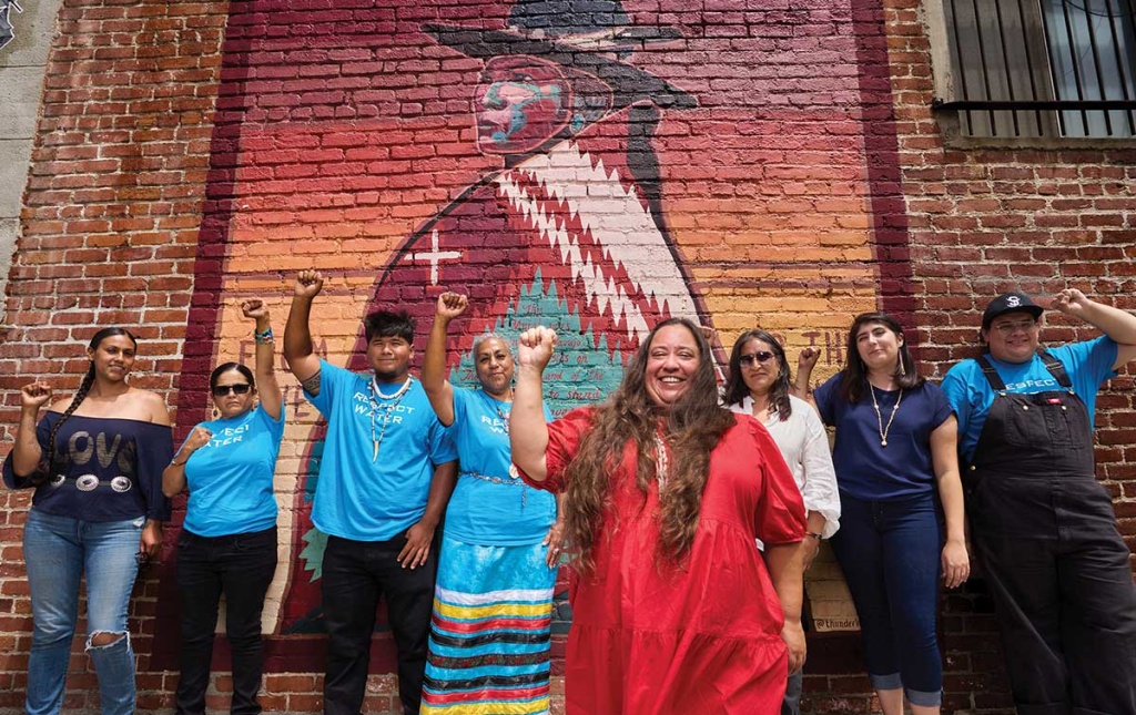 Image of Angela Mooney D'Arcy and staff holding their fists up in front of a mural on a brick wall.