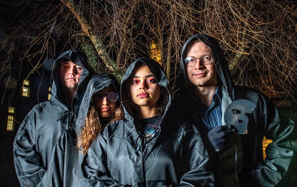 Image of four students on Brown campus dressed in black cloaks with one holding a mask