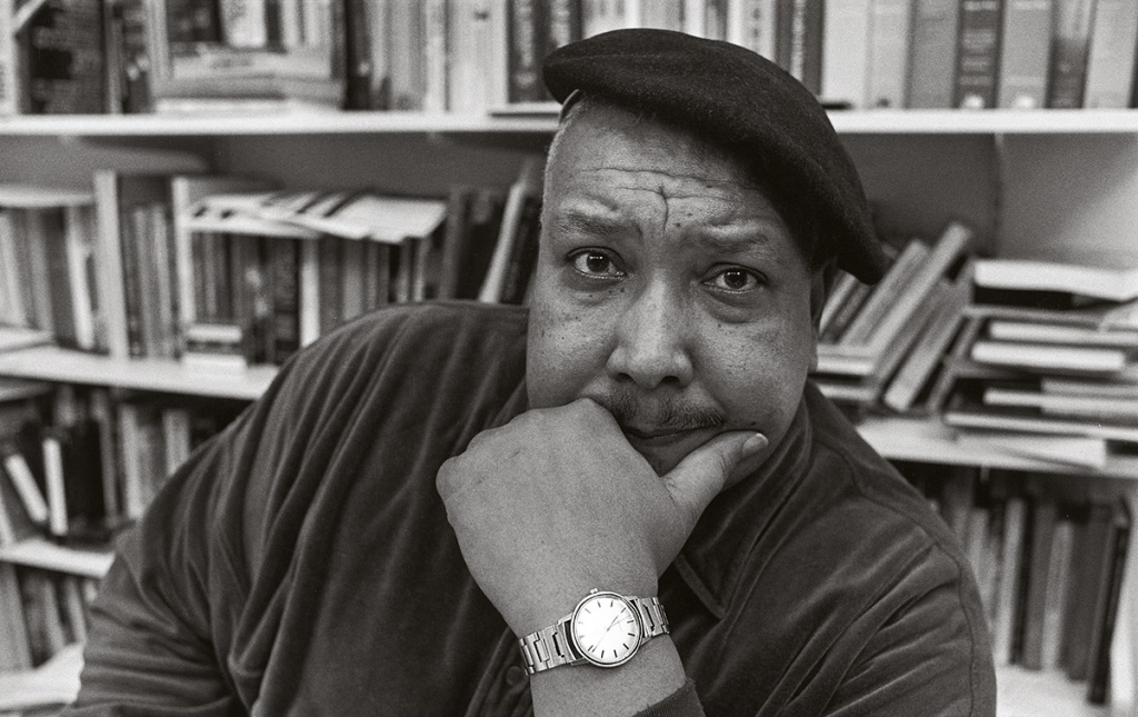 Archival image of Michael S. Harper with a beret on his head and books behind him.