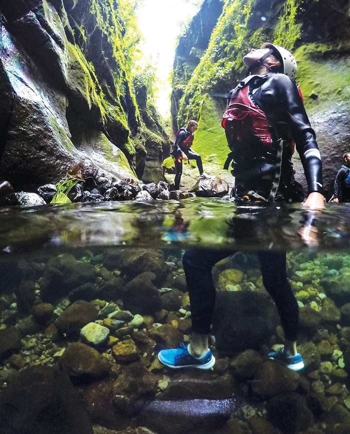 Photograph of a man standing in a river in a canyon, dressed in outdoor gear.