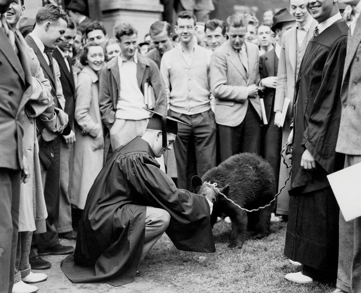 Vintage photograph of a bear being petted by a man in a cap and gown, surrounded closely by a group of people.