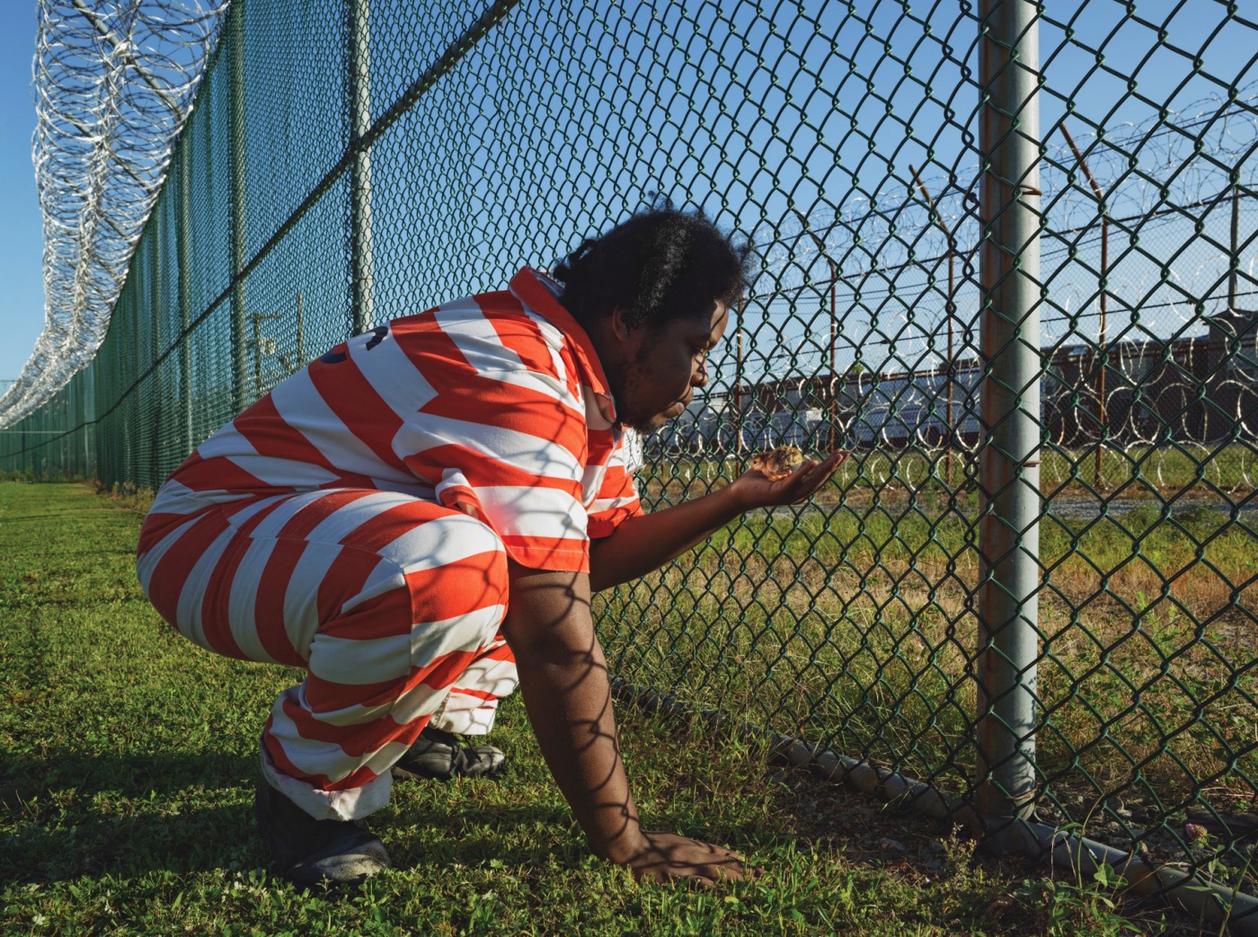 A man wearing a prison uniform squats by a fence, holding a chick in his hand.