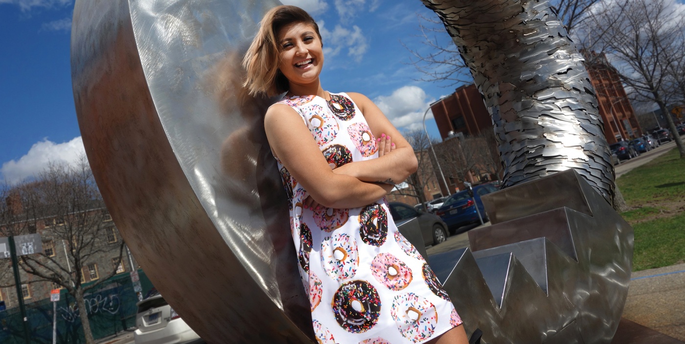 Photo of Rachael Cholak ’18 wearing a dress printed with donuts, in front of a large sculpture shaped like a donut.