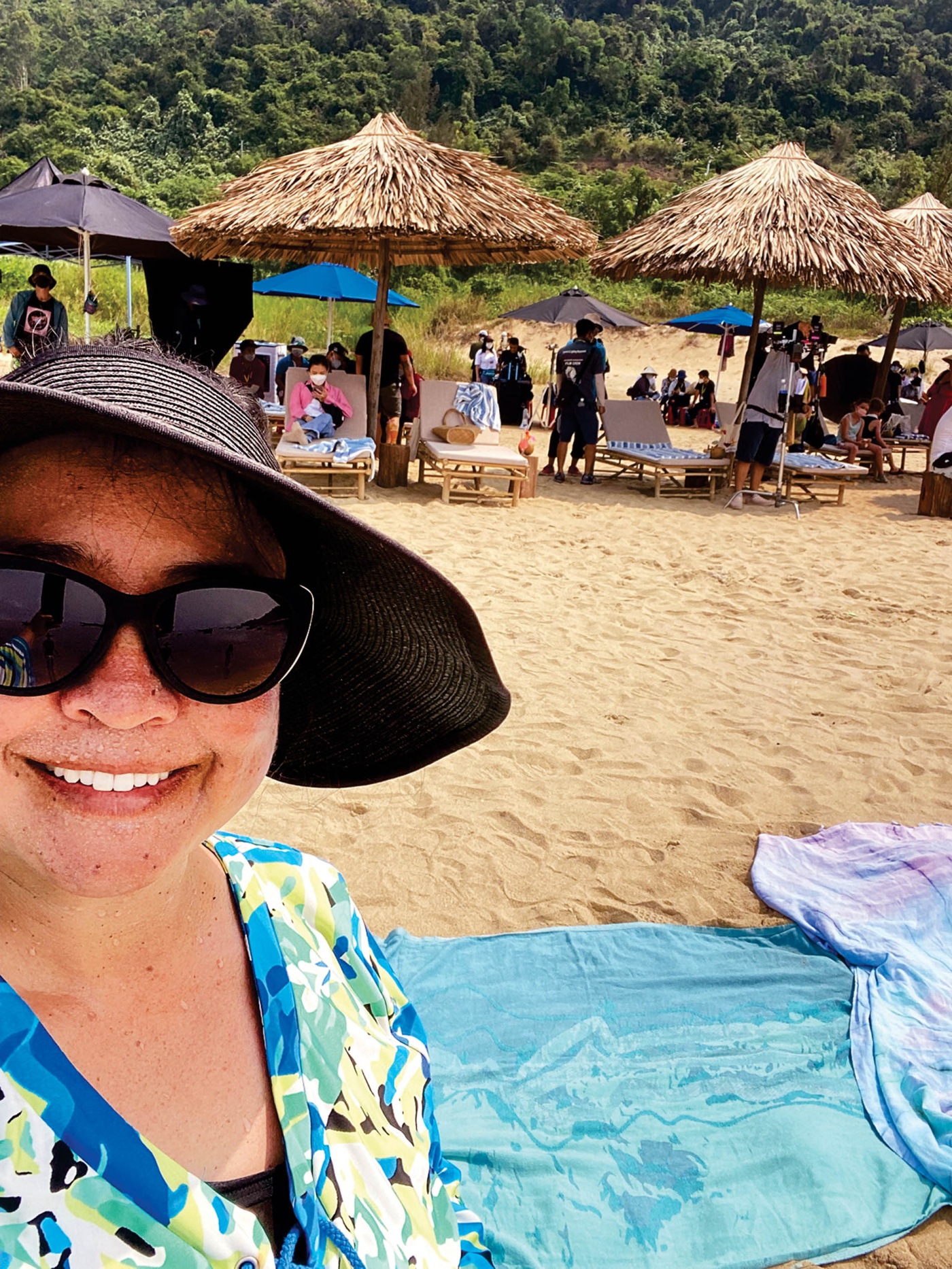 A selfie image of Eirene Donohue on set on a beach in Da Nang, Vietnam, with beach huts and people in the background.