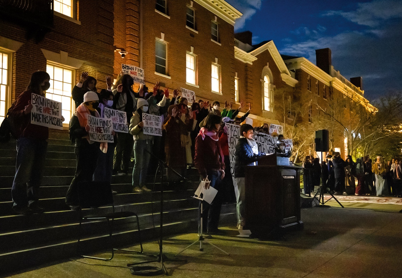 Image of students holding up signs and protesting at night with a student in front of a podium speaking in front of University Hall at Brown University