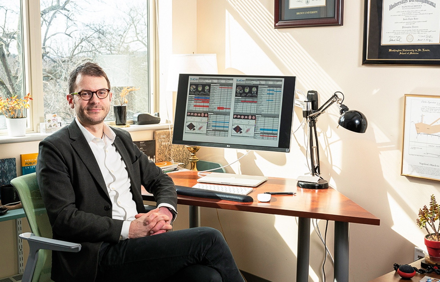 An image of Justin Baker sitting at his desk with a monitor and window behind him.