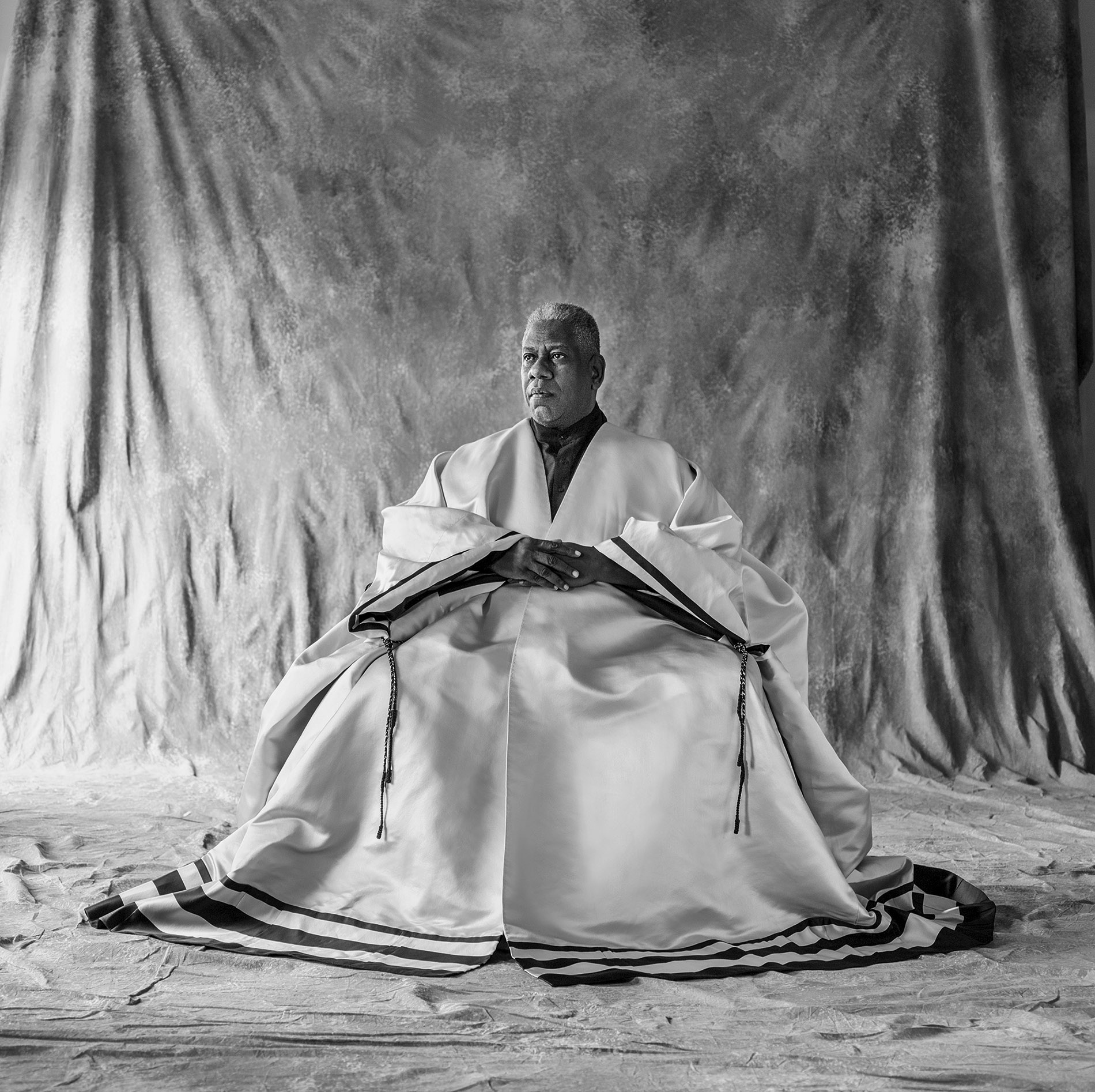Black and white image of Leon Talley sitting in a chair with his hands on his lap and fabric surrounding him