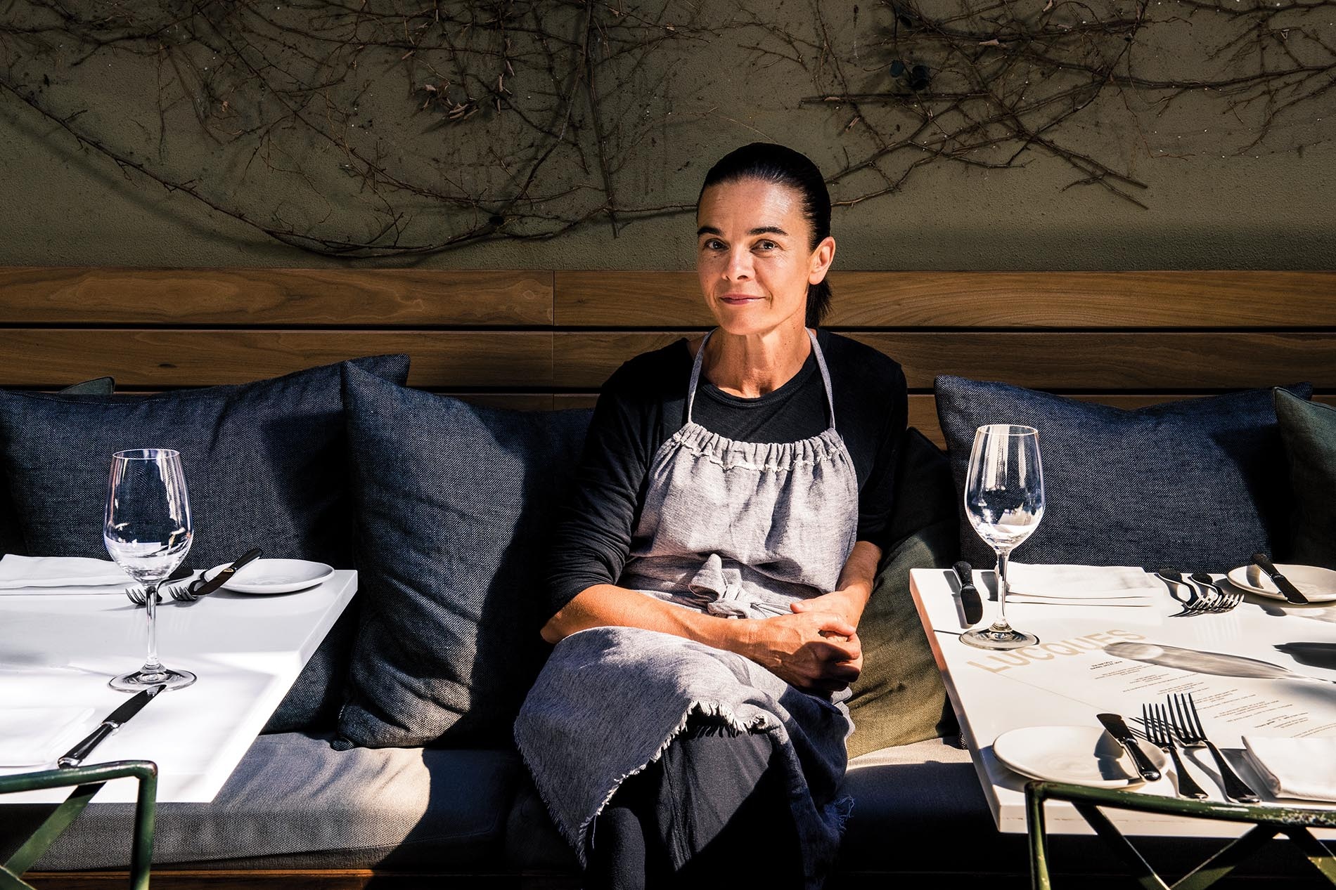 Photo of Suzanne Goin by Julia Stotz sitting on a bench behind a table at her restaurant.