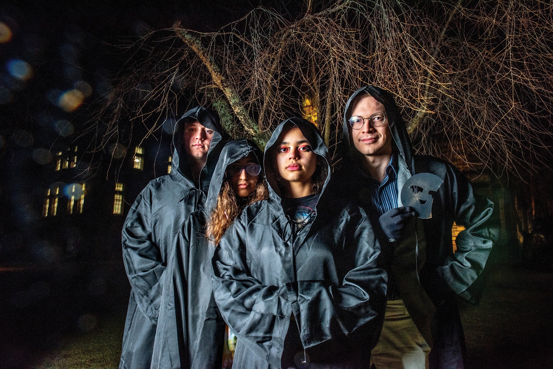 Image of four students on Brown campus dressed in black cloaks with one holding a mask