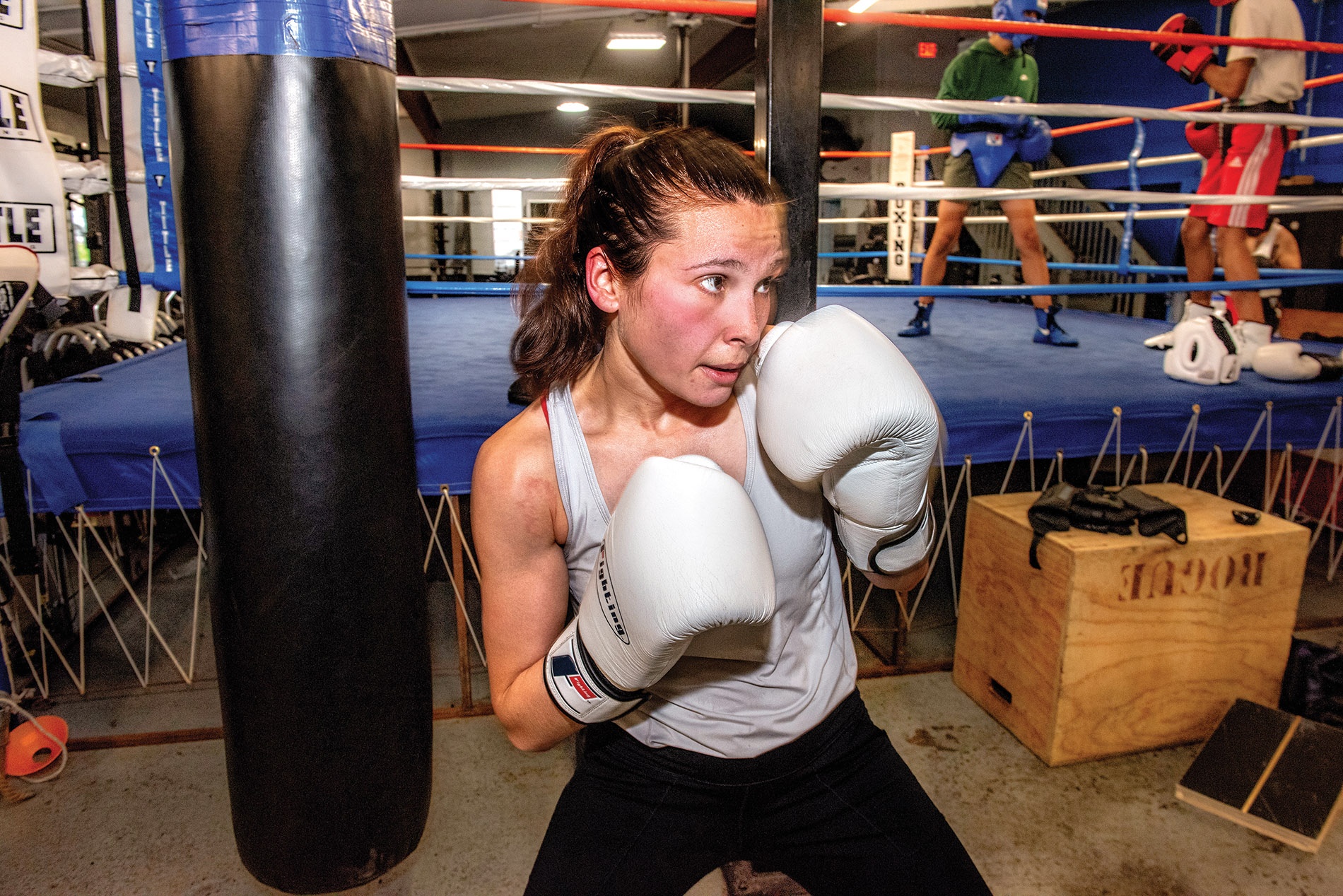 Image of Hannah Doyle holding her hands in a fighting stance with boxing gloves on them and a boxing ring in the background.