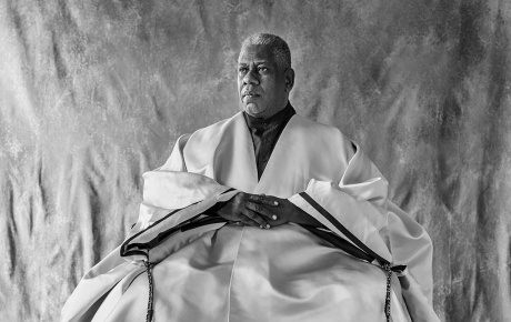 Black and white image of Leon Talley sitting in a chair with his hands on his lap and fabric surrounding him