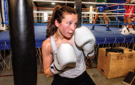 Image of Hannah Doyle holding her hands in a fighting stance with boxing gloves on them and a boxing ring in the background.