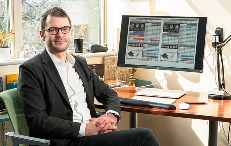 An image of Justin Baker sitting at his desk with a monitor and window behind him.